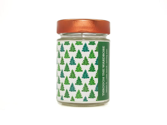 Onset & Rime balsam scented candle called "Through the Wardrobe" in a 10 oz glass jar with copper lid. The label has a snow-white background with a pattern of green fir trees and a single light post. The text on the label is "Through the Wardrobe - Balsam Fir, Cedar Boards, Swirling Snow".