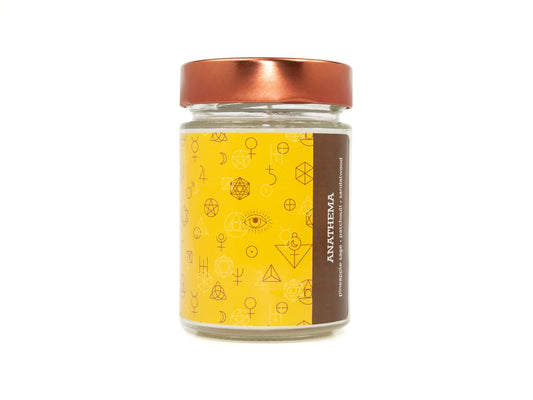 Onset & Rime earthy, herbal scented candle called "Anathema" in a 10 oz glass jar with copper lid. The label is yellow with a pattern of dark brown occult symbols . The text on the label is "Anathema - Pineapple Sage, Patchouli, Sandalwood".