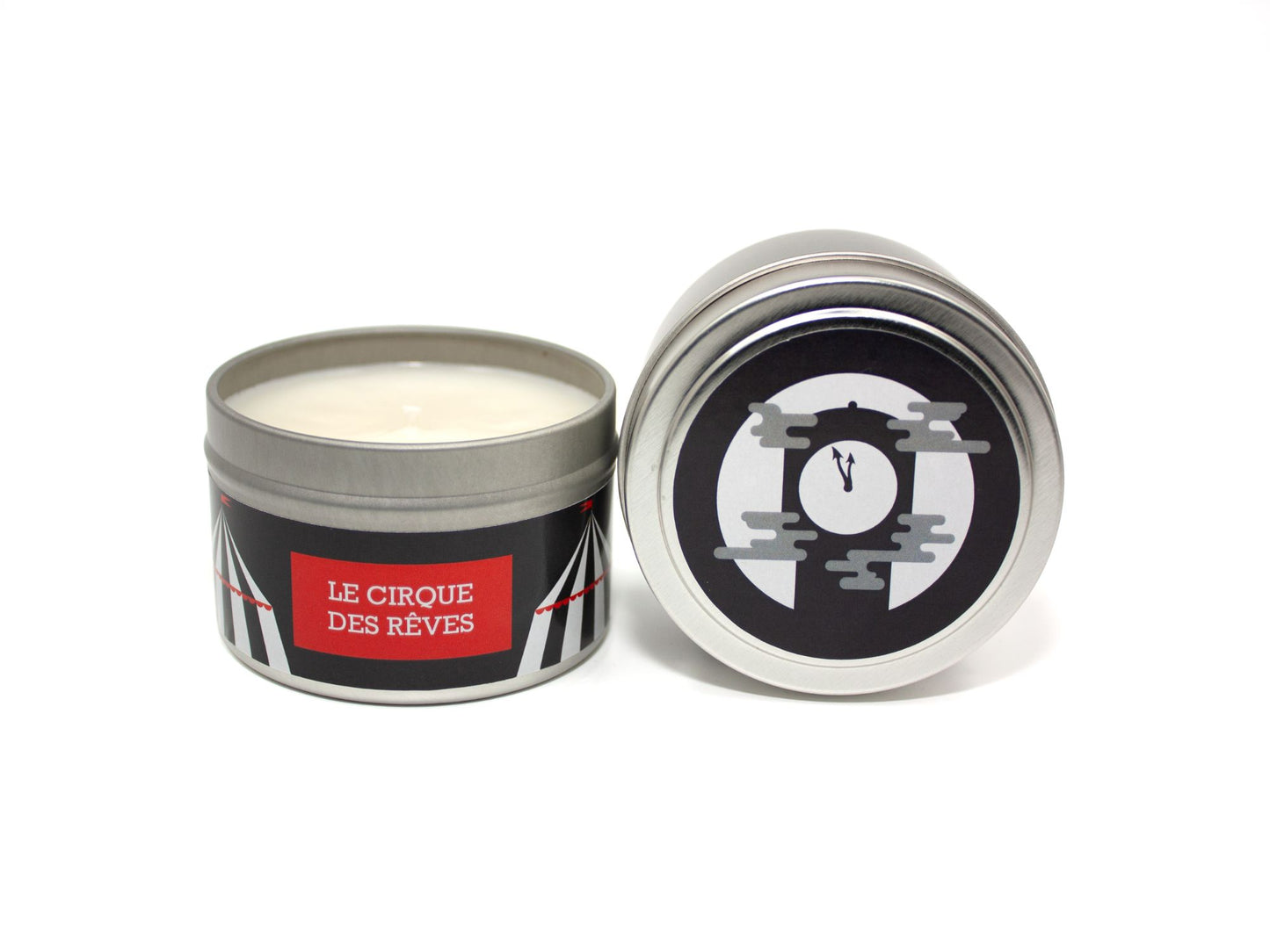 Onset & Rime caramel apple and fall air scented candle called "Le Cirque des Rêves" in a 4 oz silver tin. The circular label on top has a black background with black and white circus tents and a clock in front of a full moon. The text on the front label is "Le Cirque des Rêves - Apple, Caramel, Fallen Leaves, Bonfire".