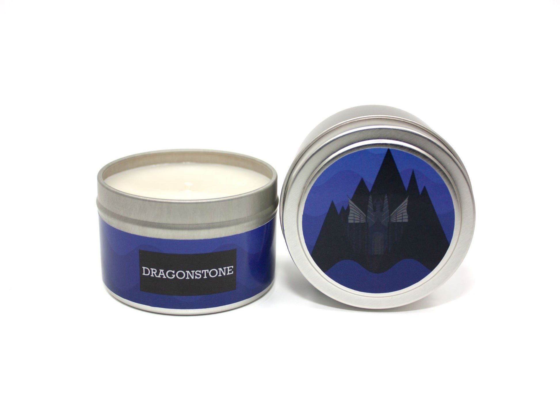 Onset & Rime chilly island scented candle called "Dragonstone" in a 4 oz silver tin. The circular label on top has blue waves in the background with a volcanic island and castle. The text on the front label is "Dragonstone - Patchouli, Vetiver, Crashing Waves, Salty Air".