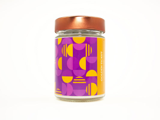 Onset & Rime chocolate bar scented candle called "Golden Ticket" in a 10 oz glass jar with copper lid. The label is a geometric pattern of purple and gold half circles. The text on the label is "Golden Ticket - Chocolate, Marshmallow Cream, Toasted Hazelnut".