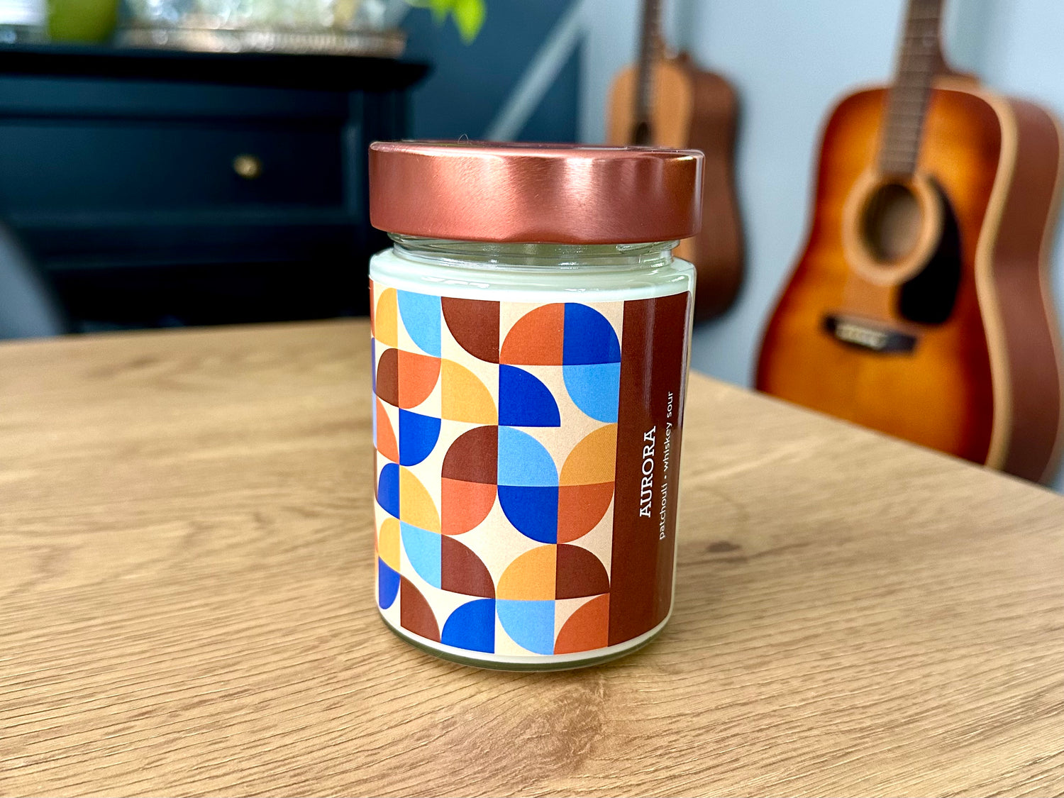 Onset & Rime rain scented candle called “Aurora” in a 10 oz glass jar with copper lid. The label is beige with a pattern of orange, yellow, blue and brown quarter circles. The text on the label is “Aurora - Patchouli, Whiskey Sour”.