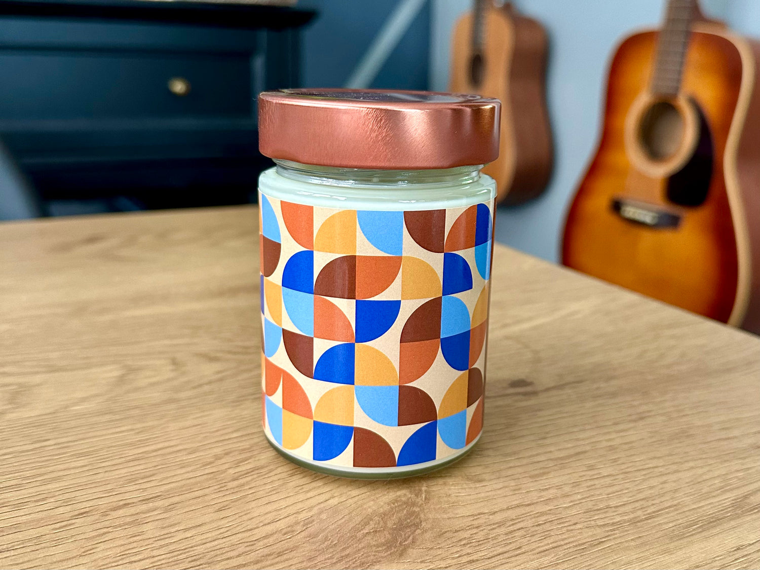 Onset & Rime rain scented candle called “Aurora” in a 10 oz glass jar with copper lid. The label is beige with a pattern of orange, yellow, blue and brown quarter circles. The text on the label is “Aurora - Patchouli, Whiskey Sour”.