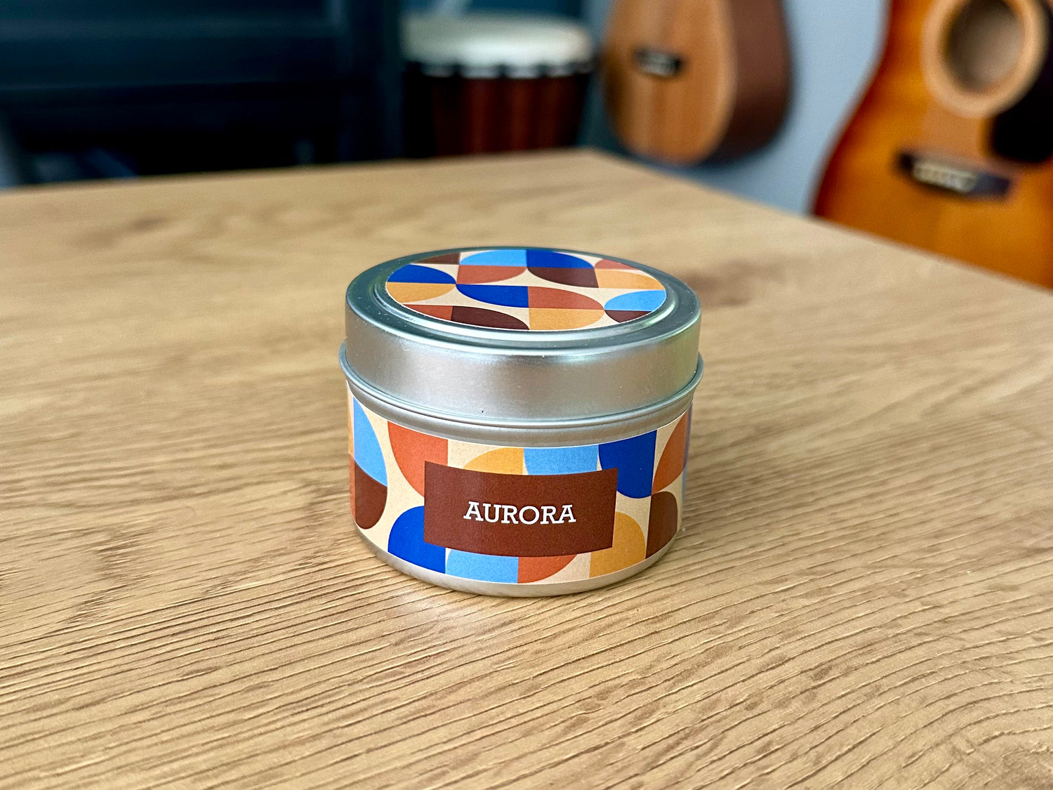 Onset & Rime rain scented candle called "Aurora" in a 4 oz silver tin. The label is beige with a pattern of orange, yellow, blue and brown quarter circles. The text on the label is “Aurora - Patchouli, Whiskey Sour”.