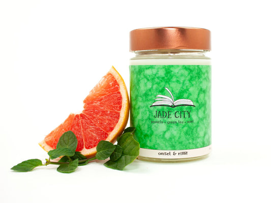 A staged photo of Onset & Rime pomelo, green tea and mint scented candle called "Jade City". The candle stands next to a slice of pomelo and sprig of mint in front of a white background.