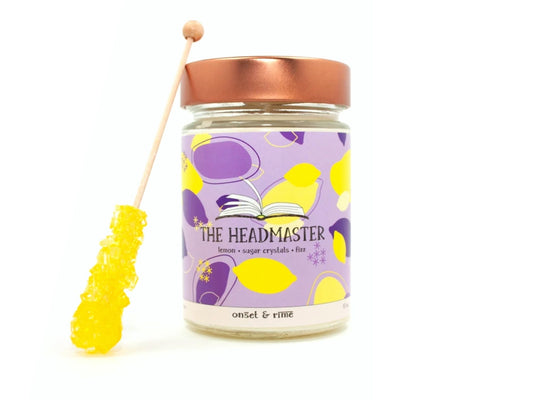 A staged photo of Onset & Rime lemon candy scented candle called "The Headmaster". The candle stands next to a lemon rock candy stick in front of a white background.