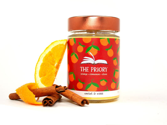 A staged photo of Onset & Rime spiced orange scented candle called "The Priory". The candle stands next to a slice of orange and some cinnamon sticks in front of a white background.